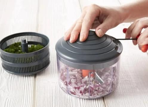 Eco-Chop manual food chopper with diced red onion inside clear bowl and removable herb basket beside it on a white wooden surface.