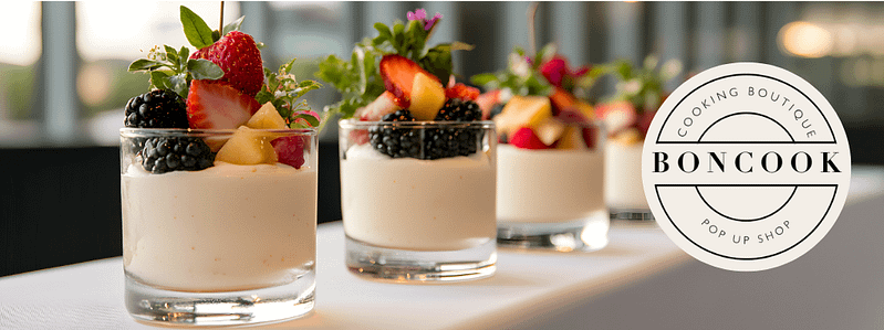 A row of six glass cups filled with creamy dessert topped with fresh berries and mint leaves, placed on a white surface with a blurred background of windows and lights.