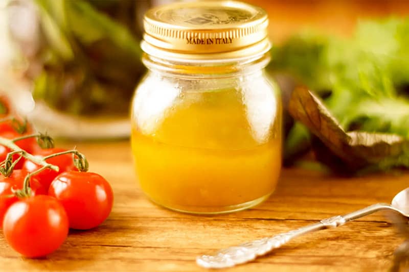Euro Vinaigrette in a glass jar with gold lid on a wooden table, surrounded by cherry tomatoes and fresh greens.